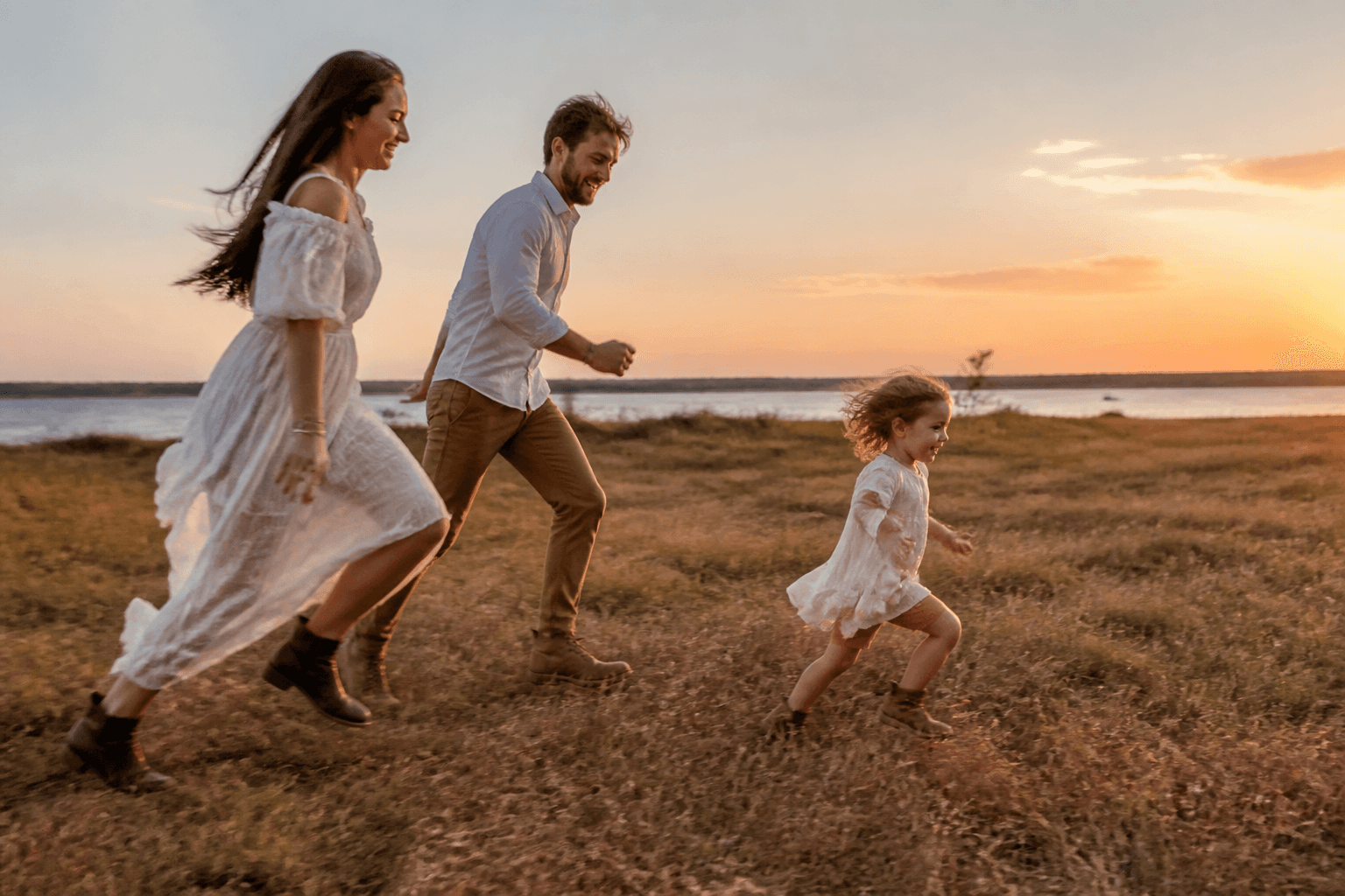 Parents and child running playfully across open grass at sunset during a candid session
